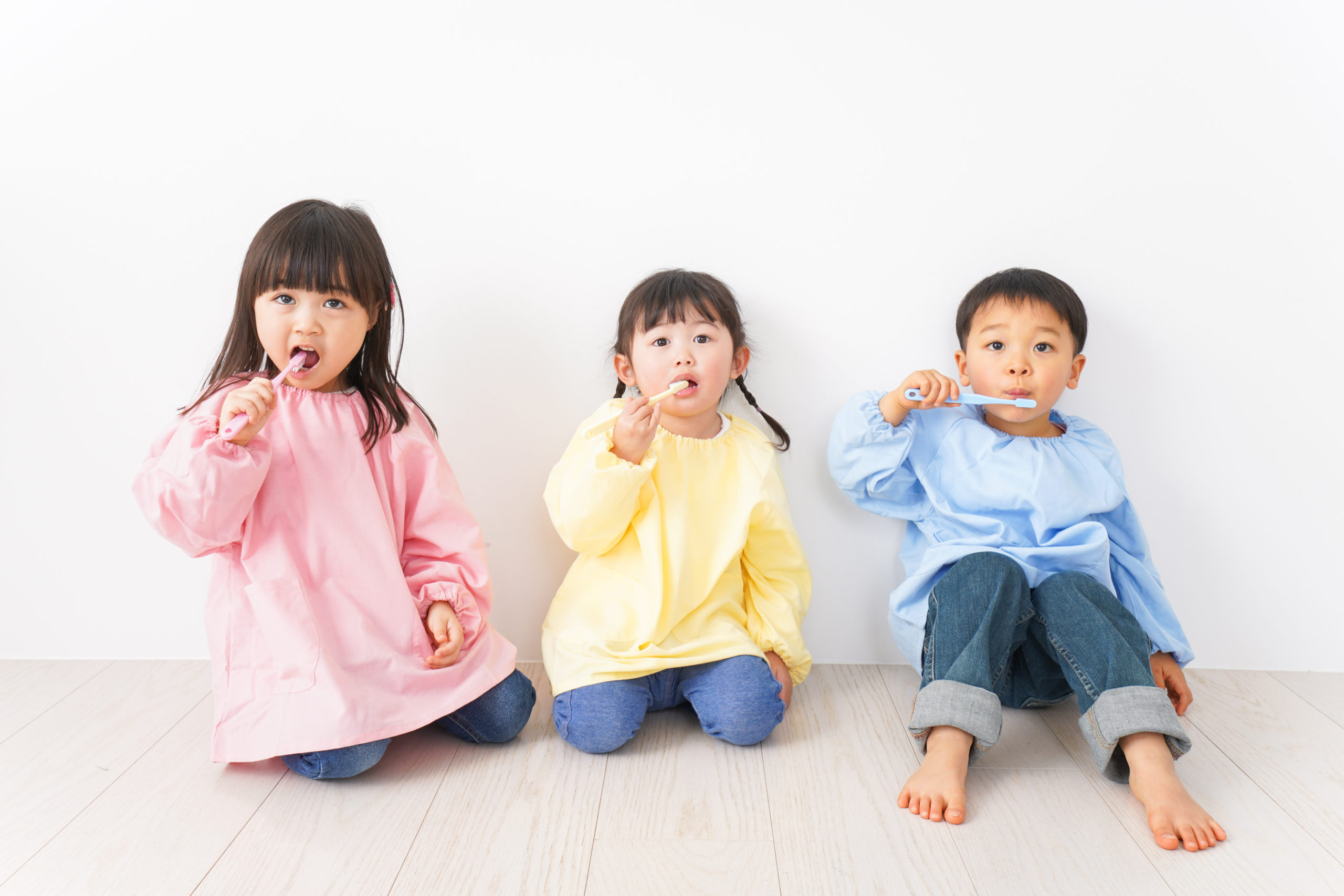 ピンクと黄色と青色の服を着たこども3人が座って歯ブラシをしている